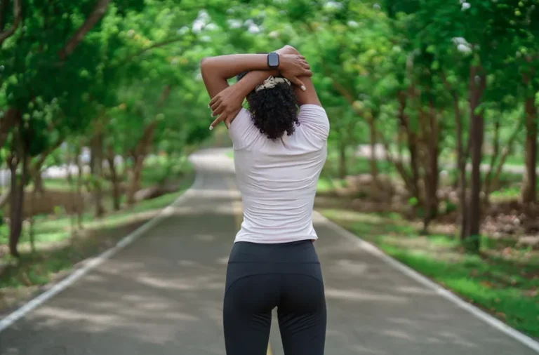 Reset Movement Routine Spring - woman standing centre of image facing away from the camera, and stretching. She is standing on a road on a spring day.